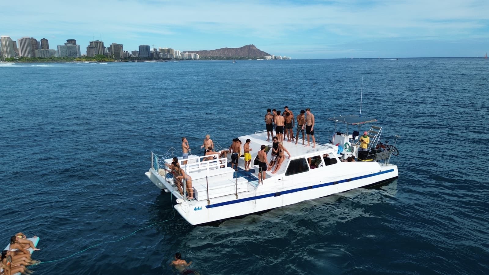 Aerial view of the Aukai catamaran anchored off Waikiki with a large group of guests on the rooftop deck and Diamond Head in the background.