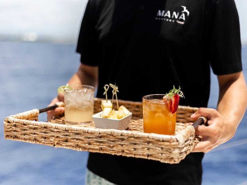 A Mana host carrying fresh cocktails and pineapple on a woven tray.